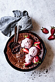 Ein Brownie aus der Pfanne, belegt mit einer Kugel Erdbeereis und frischen Erdbeeren, serviert mit zwei Löffeln auf einer strukturierten Oberfläche.