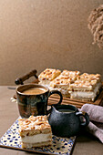 Homemade Norwegian cake Verdens Beste Kake with pudding, meringue and almonds, cut into squares, served on a wooden tray. Beige-coloured table with coffee cup, spoon, milk jug, dry reed in a bottle, beige-coloured wall.