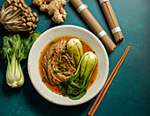 Bowl of soba noodles in spicy broth with baby bok choy, surrounded by fresh mushrooms, ginger