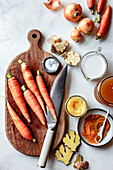 A selection of fresh ingredients, including carrots, onions and ginger, arranged on a wooden chopping board with kitchen utensils and spices.