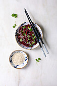 Korean-style vegan beetroot salad. Marinated grated beetroot with sesame seeds and coriander leaves in a ceramic bowl. Next to it, bowl with sesame seeds and chopsticks on a white marble base. View from above