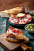 Sliced Spanish ham Jamon Iberico paleta on a black plate. Bowl of green olives, fresh and toasted white bread, olive oil, glass of red wine, tapas. Old wooden background, dark turquoise colour. Place to copy
