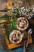 Classic Italian tiramisu, layered with cocoa powder and almonds in two glasses. Served on a tray with spoons on an old wooden table. Decorated with white baby's breath flowers and green leaves.