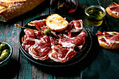 Sliced Spanish ham Jamon Iberico paleta on a black plate. Bowl of green olives, fresh and toasted white bread, olive oil, glass of red wine, tapas. Old wooden background, dark turquoise colour. Place to copy