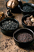 Set of five black spices: black fermented garlic, black Himalayan salt, black mustard seeds, black cardamom and fermented campota pepper in black ceramic bowls. Dark wooden background. Close-up