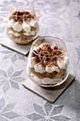 Classic Italian tiramisu, served in two glasses. Layers of coffee-soaked sponge cake and creamy mascarpone, dusted with cocoa powder. Placed on a patterned grey-blue ceramic tile, spoon next to it