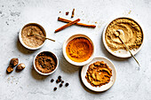 A selection of spices in bowls with small spoons on a textured surface, with cinnamon sticks, nutmeg and allspice.