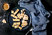 Traditional Spanish dessert classic: soft orange turron and crunchy almond turron, cut into pieces on a blue ceramic plate. Linen napkin, espresso coffee in black cup, black wooden background, top view.