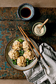 Homemade Asian steamed dumplings in a grey plate, topped with sesame seeds and green onions. Soy sauce, chopsticks, grey napkin nearby on a rustic wooden board, beige textured background.