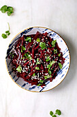 Korean-style vegan beetroot salad. Marinated grated beetroot with sesame seeds and coriander leaves in a ceramic bowl. Next to it, bowl with sesame seeds and chopsticks on a white marble base. View from above