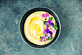 A beautifully presented bowl of creamy hummus drizzled with olive oil and decorated with vibrant edible flowers, served on a textured blue background. A modern interpretation of a classic dish.