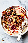 Eine Schale Joghurt mit Müsli, Pekannüssen, Kürbiskernen, Granatapfelkernen und Apfelspalten auf einer Marmorplatte.