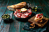 Sliced Spanish ham Jamon Iberico paleta on a black plate. Bowl of green olives, fresh and toasted white bread, olive oil, glass of red wine, tapas. Old wooden background, dark turquoise colour. Place to copy
