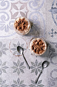 Classic Italian tiramisu, served in two glasses. Layers of coffee-soaked sponge cake and creamy mascarpone, dusted with cocoa powder. Placed on a patterned grey-blue ceramic tile, spoon next to it