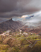 Aerial view of roque bentayga with picturesque village and winding road amidst dramatic sky and clouds, Tejeda, Las Palmas, Gran Canaria, Spain.