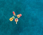 Aerial view of women in bikinis holding hands on inflatable mattresses on Atokos island, Greece.