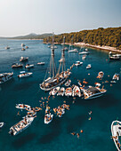 Aerial view of crowded Otok Koludarc with transparent turquoise water and leisure vessels, Primorje-Gorski Kotar, Croatia.