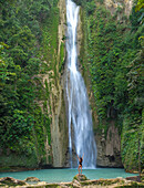 Aerial view of majestic mantayupan falls with a woman standing on a rock surrounded by lush tropical forest, Campangga, Barili, Cebu, Philippines.