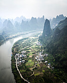 Aerial view of beautiful karst mountains and a serene river winding through a lush valley with a picturesque village, Guilin, China.