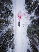 Aerial view of a man lighting a pink smoke grenade on a snowy road in the forest in Estonia.