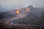 Aerial view of lava flowing through the melting rocks on mountain top with magma sprinkling from craters and smoke all around, Mýrdalshreppur, Iceland.