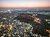 Luftaufnahme des Mehrangarh Forts, beleuchtet in der Abenddämmerung mit pulsierendem Stadtbild und Abendhimmel, Jodhpur, Indien.