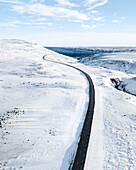 Drohnenaufnahme einer Autobahn, die sich durch eine schneebedeckte Landschaft im Norden Islands schlängelt.