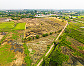 Luftaufnahme einer abwechslungsreichen Landschaft mit unbefestigter Straße, teilweise abgebranntem Ackerland und Bäumen, Phra Non, Nakhon Luang, Ayutthaya, Thailand.