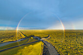 Aerial view of full rainbow over green landscape with road, river and bridge, south Iceland