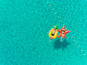 Aerial view of man and woman floating by string buoy on inflatable mattresses in transparent blue sea.