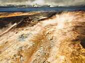 Aerial view of a volcanic hot spring with sulphuric smoke and a lake in background, Namafjall Hverir viewpoint, Northeastern Region, Iceland.