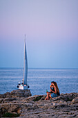 Couple enjoying sunset views on Pendma Chiatt (Pietra Piatta) with sailing yacht in the background, Polignano a Mare, Puglia, Italy