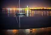 A sailboat rests in calm waters as Isla Cristina's lights twinkle in the distance, while a cat strolls along the shore at night.
