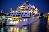 A large cruise ship is moored on the Guadalquivir River in Seville, illuminated at night, showcasing the vibrant atmosphere of the city.