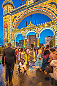 Seville, Spain, Apr 19 2018, Visitors gather near the brilliantly lit entrance gate of the April Fair as vibrant festivities unfold at twilight in Seville.