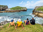Friends sit on grassy cliffs, looking out over sea stacks and turquoise waters along the rugged cliffs of Asturias in Spain.