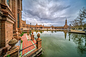 Visitors admire the intricate design of Plaza de España in Seville, reflecting in the serene water while clouds drift overhead.
