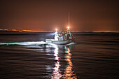 A fishing boat navigates the calm waters of Punta del Moral, illuminated by lights as it heads out for an evening catch under a dark sky.