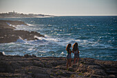 Zwei Mädchen, die auf dem Pendma Chiatt (Pietra Piatta) spazieren gehen, während im Hintergrund die Wellen an die Felsen schlagen, Polignano a Mare, Apulien, Italien