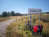 A cyclist arrives at Cubillos, Soria, with red panniers resting beside the sign, enjoying the quiet of forgotten ruins in summer.