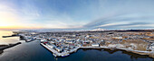 Aerial view of the town's harbor embraced by the cold sea, houses dusted with snow, under a sky painted with dawn's first light, Grindavik, Grindavíkurbær, Iceland.