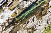 Aerial view of crystal-clear turquoise water cascading through rugged rocks where sunbathers relax in the sun-drenched Verzasca River, Verzasca, Ticino, Switzerland.