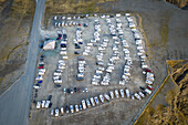 Luftaufnahme eines Parkplatzes voller Autos und Wohnmobile. Die Landschaft ist in gedämpften Braun- und Grautönen gehalten, die im Kontrast zu den bunten Fahrzeugen stehen und den Overtourism vom Feinsten zeigen, Nordkapp, Finnmark, Norwegen.