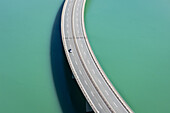 Luftaufnahme eines einsamen Autos, das die Brücke über das leuchtend türkisfarbene Wasser der Drau überquert, ein eindrucksvoller Kontrast zwischen von Menschenhand geschaffenen Strukturen und der Schönheit der Natur, Ferlach, Kärnten, Österreich.