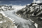 Luftaufnahme des Rhonegletschers, der sich unter einem weiten Himmel durch zerklüftete, schneebedeckte Gipfel schlängelt, eine Symphonie aus grauem Eis vor einem strahlend weißen Hintergrund, Obergoms, Wallis, Schweiz.