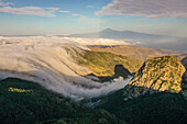 Luftaufnahme der zerklüfteten, grünen Landschaft, in der sich die Wolken wie ein sanfter, weißer Wasserfall über die Berggipfel ergießen, La Gomera, Kanarische Inseln, Spanien.