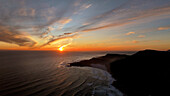 Aerial view of sunset over serene waves and majestic mountains along the coastal shoreline, Scarborough, Western Cape, South Africa.