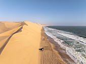 Luftaufnahme von schönen Sanddünen, die auf den Ozean treffen, mit ruhigen Wellen und Fahrzeugspuren, Sandwich Harbour, Erongo, Namibia.
