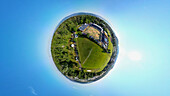 Aerial view of a tiny planet perspective featuring a scenic urban landscape with a soccer field, stadium, and residential buildings surrounded by greenery, Lyss, Bern, Switzerland.