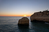 Aerial view of serene sunset over rugged cliff beach with rocks and tranquil ocean, Algarve, Portugal.